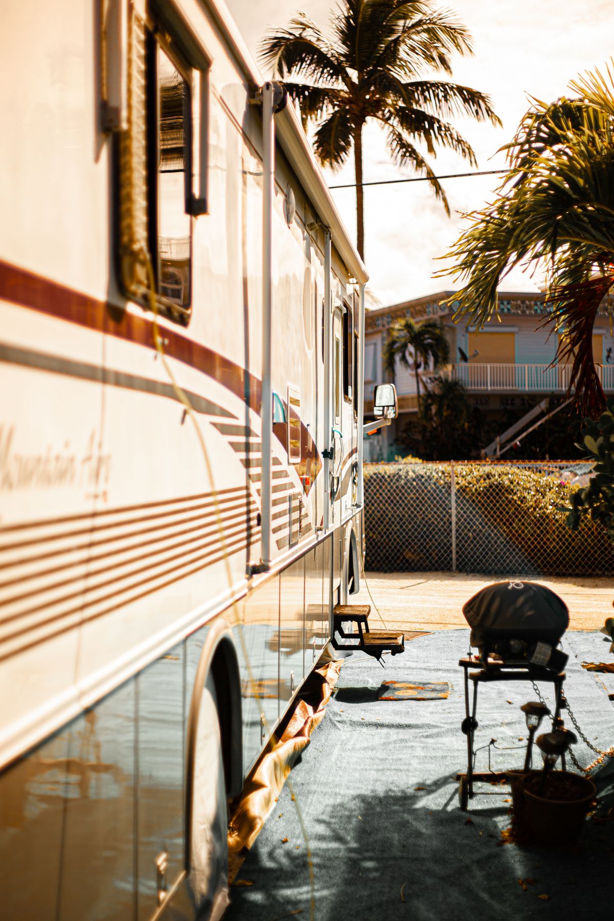 Side of a motorhome parked at an RV site with palm trees and a barbecue grill nearby